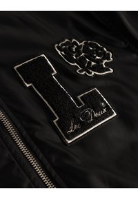 Must mustriga bomber-jakk mustast kangast, millel on tekstuuriga must "L" aplikatsioon ja lilleline tikand. Omab hõbedast tõmblukku ja valget "Les Deux" logo.