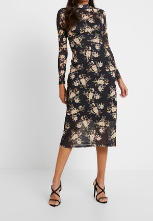 Vrouw draagt een zwarte midi-jurk met lange mouwen en beige bloemenpatronen, gecombineerd met zwarte sandalen met hoge hakken en bandjes, staand tegen een witte muur.