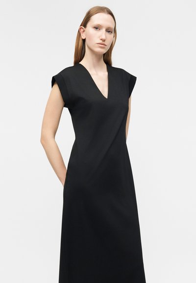 Γυναίκα με ίσια ανοιχτό καστανά μαλλιά που φοράει ένα αμάνικο μαύρο φόρεμα με ντεκολτέ V, στέκεται μπροστά σε ένα απλό φωτεινό φόντο.