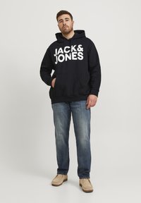 Marineblauer Kapuzenpullover mit großem weißen "JACK & JONES"-Logo, frontaler Kängurutasche und Rippbündchen. Getragen mit blauen Jeans und beigen Schuhen.