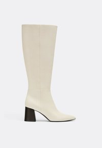Botte haute en cuir crème avec bout pointu et talon bloc épais marron foncé, vue de profil sur un fond clair.