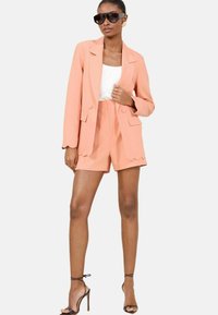 Peach blazer with scalloped hem and matching shorts; white inner top; paired with dark oversized sunglasses and strappy high heels.