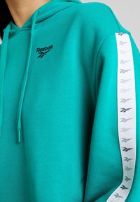 Haut à capuche turquoise avec cordon de serrage et logo Reebok noir. Présente une rayure latérale blanche avec des logos gris répétés et un tissu doux et texturé.