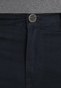 Gros plan sur la ceinture d’un jean bleu foncé avec un bouton argenté gravé « PME Legend Jeans » et une partie d’un pull en maille gris au-dessus.