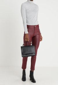 Persona vestida con un suéter de cuello alto gris claro y pantalones de cuero burdeos, sosteniendo un bolso negro y usando botas negras hasta el tobillo, sobre fondo blanco.