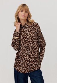 Kobieta z blond włosami, ubrana w brązową koszulę w trędy lamparta z guzikami oraz ciemnoniebieskie spodnie, pozująca z ręką przy szyi na jednolitym tle.