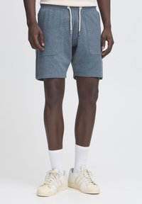 Shorts en coton bleu texturé avec une taille élastique et un cordon de serrage. Comprend deux poches latérales et une coupe décontractée. Portés avec des chaussettes blanches et des baskets.