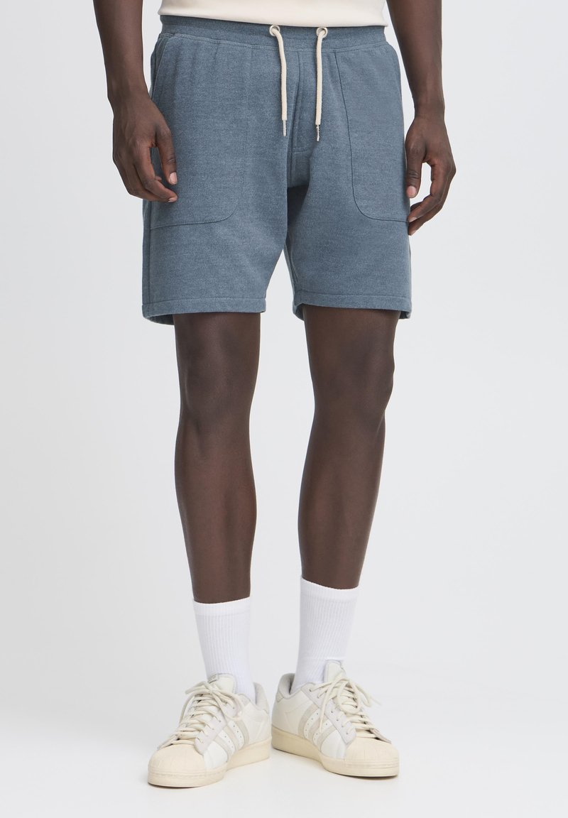 Shorts en coton bleu texturé avec une taille élastique et un cordon de serrage. Comprend deux poches latérales et une coupe décontractée. Portés avec des chaussettes blanches et des baskets.