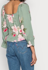 Donna che indossa una blusa floreale verde con vita arricciata e jeans denim blu, mostrata di spalle su uno sfondo semplice.