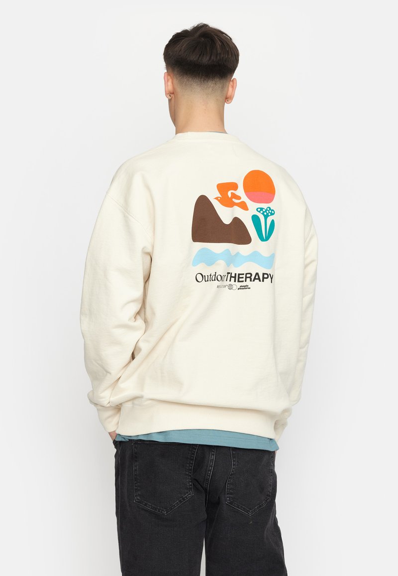 Lichtcreme sweatshirt met een grafisch ontwerp van bergen, een zon en een bloem in oranje, blauw en groene kleuren. Inclusief de tekst "OutdoorTHERAPY".