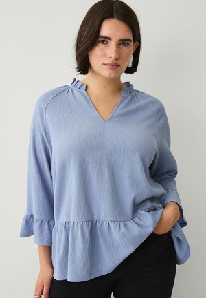 Kvinde iført en lyseblå tekstureret bluse med flæsekant ved krave og kant, 3/4-ærmer og sorte bukser, stående mod en ensfarvet baggrund.