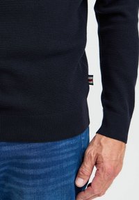 Marinefarget teksturert genser med ribbekant og mansjetter, med en liten tri-farget logo på siden. Kombinert med blå denimjeans.