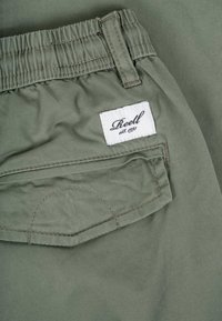 Pantalones de tela verde con cintura elástica y un bolsillo trasero que tiene una etiqueta blanca con la inscripción "Reell est. 1997" en letra negra.