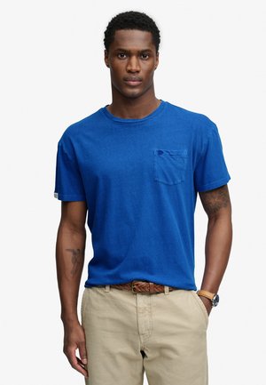 Mężczyzna z krótkimi kręconymi włosami, ubrany w niebieską koszulkę z kieszenią, beżowe spodnie, brązowy pleciony pasek, zegarek na nadgarstku oraz widoczne tatuaże na ramionach.