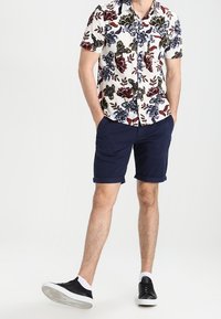 Kratší rukáv pánská košile s knoflíky v bílé barvě s vícebarevným květinovým vzorem, v kombinaci s námořnickými šortkami a černými teniskami.