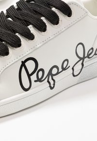 Baskets blanches avec des lacets noirs épais et le logo manuscrit « Pepe Jeans » sur le côté près de la semelle.