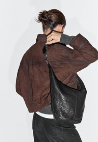 Personne avec les cheveux en chignon portant une veste marron et un pantalon noir, tenant un sac à bandoulière en cuir noir texturé derrière son dos.