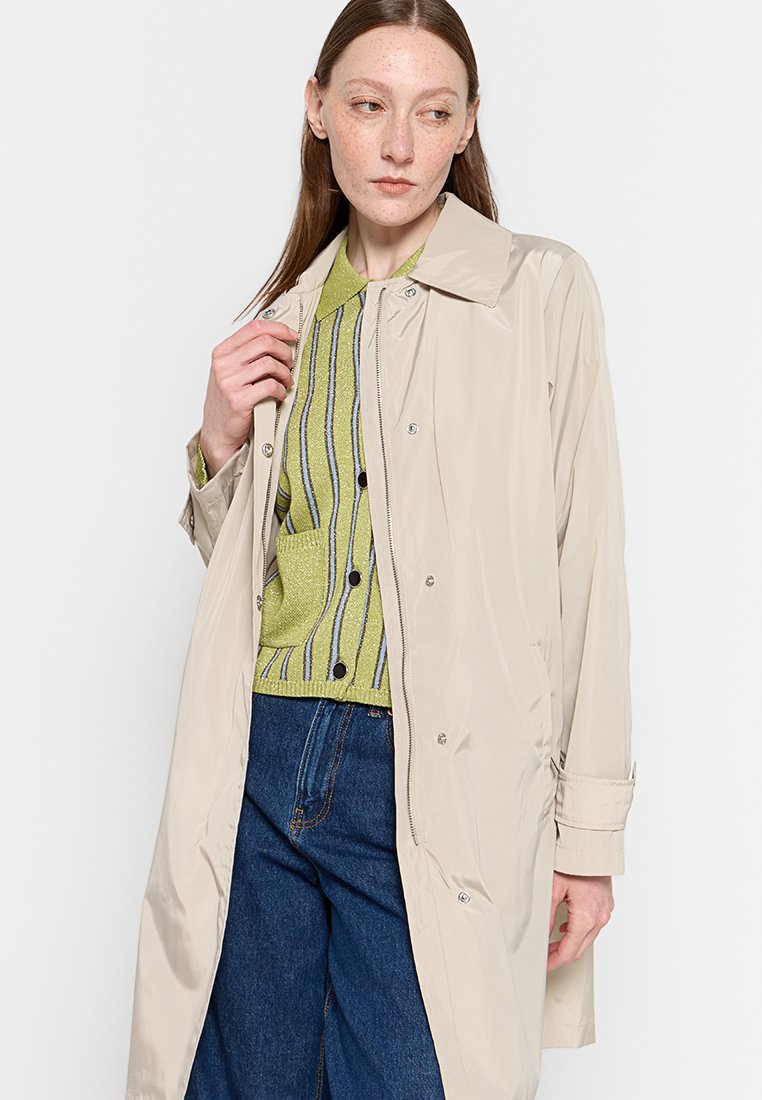Un manteau de pluie long beige avec un col, fabriqué dans un matériau brillant. Il présente des fermetures à pression et est associé à un pull à rayures vertes en dessous.