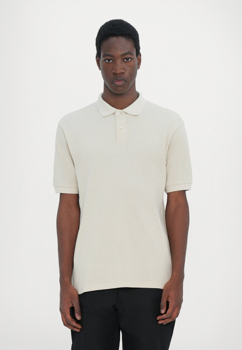 Polo beige à manches courtes, avec un col et une patte de boutonnage à trois boutons. Texture lisse avec un logo discret sur la poitrine.
