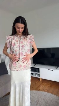 Femme en blouse fleurie et jupe blanche touchant son ventre, debout dans un salon avec une télévision et un canapé en arrière-plan.