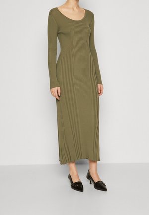 Olijfgroene ribbelige jurk met lange mouwen, een ronde halslijn en een aangesloten pasvorm, gecombineerd met zwarte pumps met puntige neus en decoratieve hardware.