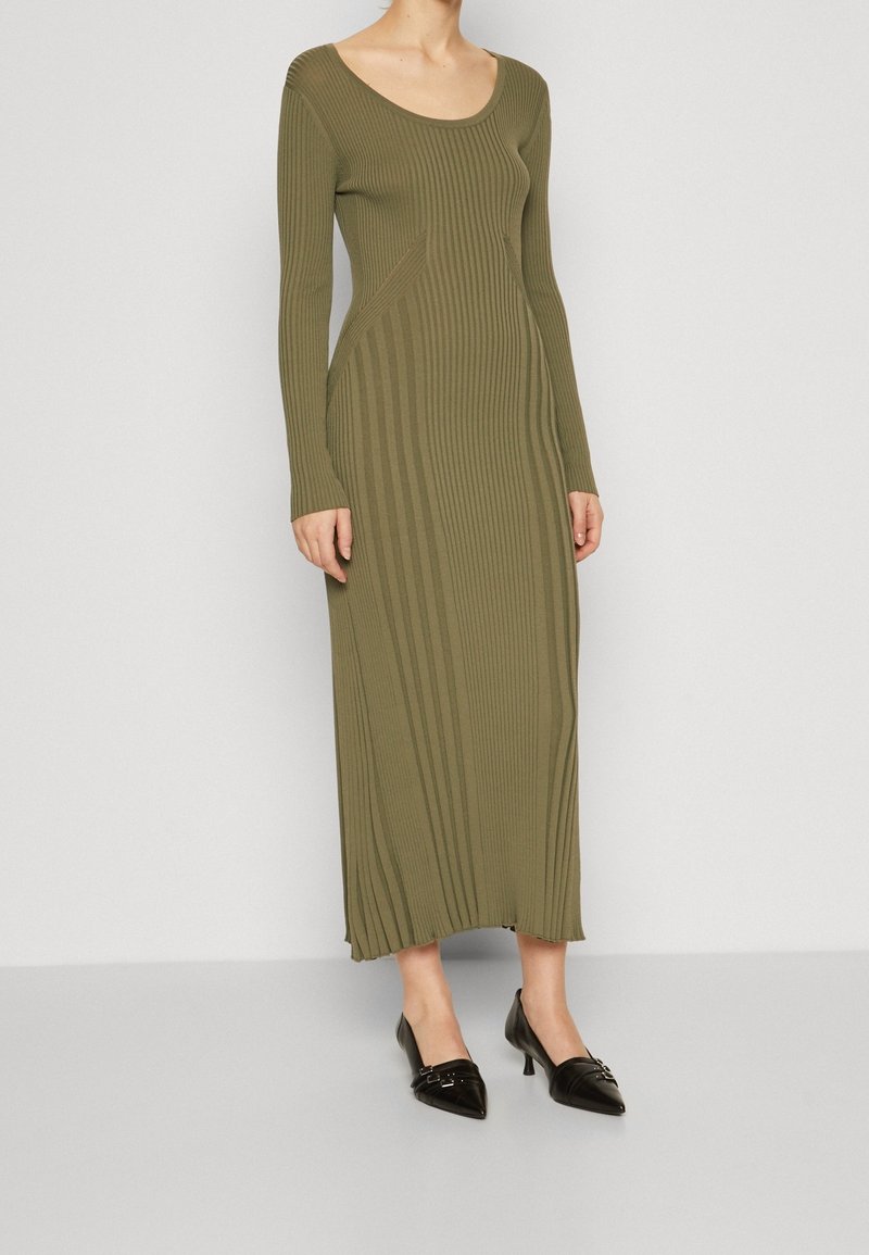 Robe côtelée à manches longues vert olive avec un col en forme de scoop et une coupe ajustée, associée à des escarpins noirs à bouts pointus ornés de ferrures décoratives.