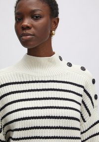 Strickpullover mit hohem, geripptem Kragen, versehen mit schwarzen horizontalen Streifen auf einem cremefarbenen Hintergrund und schwarzen Knopfakzenten an der Schulter.