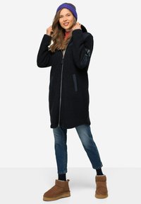 Cappotto nero foderato in pile con zip frontale, due tasche laterali e cappuccio, abbinato a jeans blu e stivali ankle marroni. Accentuato con un berretto viola.