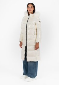 Cappotto imbottito lucido beige con cappuccio foderato in pelliccia, zip frontale e tasche laterali. Abbinato a jeans blu a gamba larga e sneakers bianche.
