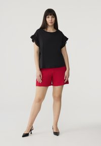 Top nero con maniche arricciate e pantaloni rossi. Il top ha una vestibilità morbida; i pantaloni sono rossi solidi con taglio dritto. Tacchi neri completano l'outfit.