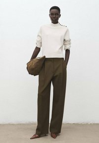 Cremfarbener Strickpullover mit hohem Kragen und Knopfdetails, kombiniert mit hochgeschnittenen olivfarbenen Hosen und einer braunen Wildlederhandtasche.