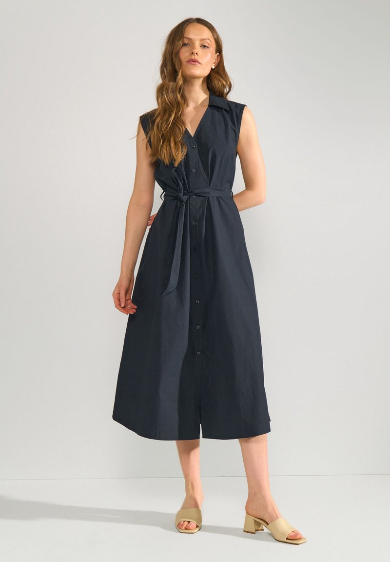 Femme aux cheveux longs portant une robe midi bleu marine sans manches avec des boutons et une ceinture nouée à la taille, ainsi que des sandales à talons beiges.