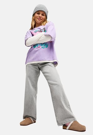 Young woman wearing a purple graphic shirt, grey wide-leg jeans, beige slip-on shoes, and a grey knit beanie, standing with arms crossed.