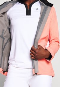 Veste extérieure légère corail avec doublure grise, poches zippées et accents en mesh, portée sur un haut blanc à manches courtes avec un col noir.