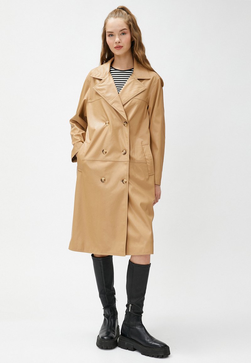 Brun faux läder trenchcoat med dubbelknäppning, stor krage, framfickor och knä-lång längd. Bärs med svarta stövlar som går upp till knäna.