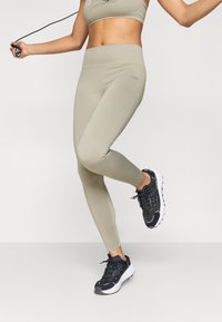 Világos bézs leggings sima textúrával, magas derékpánttal és logós részlettel. Fekete sportcipővel párosítva, fehér talppal.