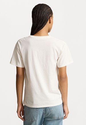 Person med fletninger, iført en enkel hvid kortærmet t-shirt og lyseblå jeans, vist bagfra mod en hvid baggrund.