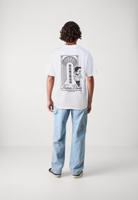 Un jeune homme aux cheveux bouclés portant un t-shirt blanc avec un graphique noir représentant un arc, du texte japonais et un personnage de dessin animé, associé à un jean bleu clair.