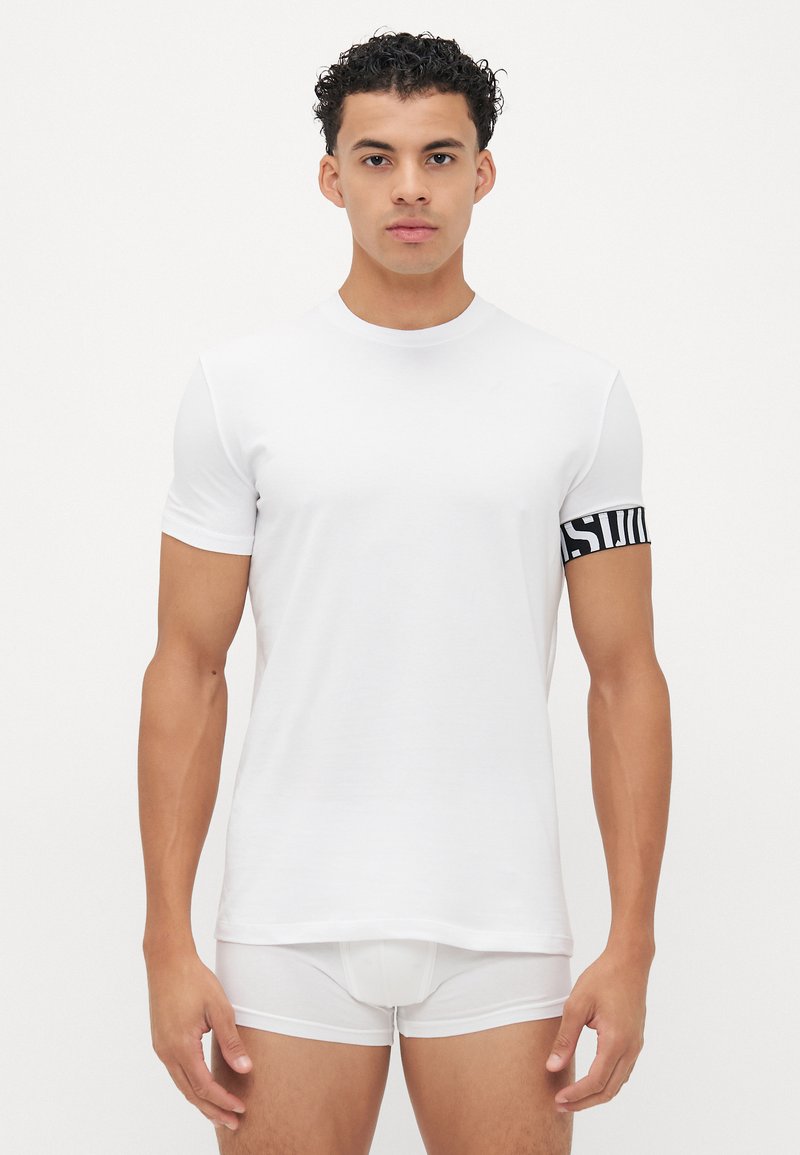 Weißes T-Shirt mit kurzen Ärmeln aus Baumwolle und rundem Ausschnitt. Verfügt über ein schwarzes Markenband am linken Arm. Getragen mit weißen Boxershorts.