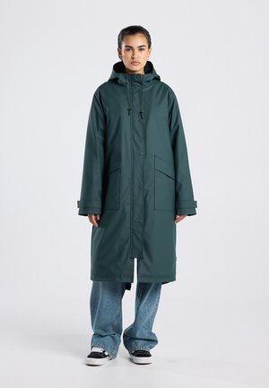 Frau trägt einen langen dunkelgrünen Kapuzen-Regenmantel, blaue Jeans und schwarz-weiße Sneakers und steht vor einem einfarbigen hellen Hintergrund.