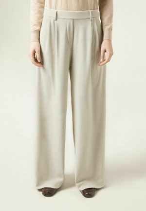 Kobieta ubrana w beżowe spodnie z wysokim stanem i szerokimi nogawkami, stojąca z rozluźnionymi ramionami wzdłuż ciała, zestawiona z ciemnymi butami o kwadratowym nosku.