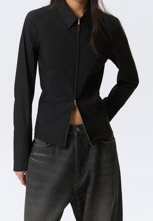 Femme portant un haut noir ajusté à manches longues avec une fermeture éclair partiellement ouverte à l'avant, associé à un jean noir ample.