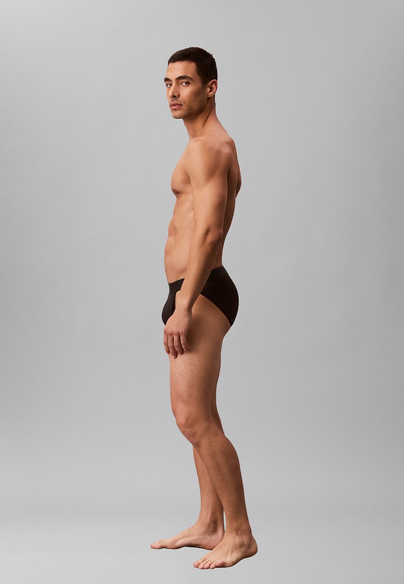 Schwarze Slips im Brief-Stil mit glatter Textur, die ein einfaches Design ohne sichtbare Markenkennzeichnung aufweisen. Modell von der Seite gesehen.