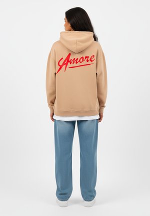 Beige oversized hoodie met een rode "Amore" graphic op de achterkant, gecombineerd met lichtblauwe wijde broek. Gladde textuur, casual stijl.