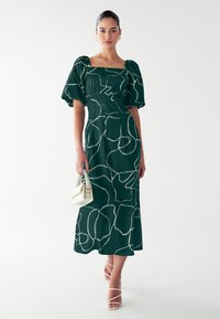 Groene jurk met witte abstracte lijnpatronen, met een vierkante halslijn en pofmouwen, gecombineerd met witte sandalen met bandjes en een kleine handtas.