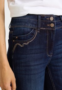 Jeans de mezclilla azul oscuro con cintura alta, costuras decorativas y cierre de dos botones. Diseño distintivo de bolsillos que realza el detalle.