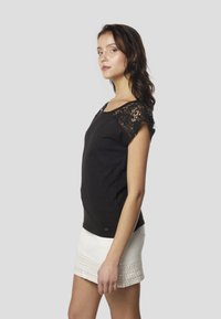 Melna īsroku krekls ar mežģīņu akcentiem uz pleciem, kopā ar baltu svārku ar dekoratīvām apmalēm. Gluda tekstūra, ikdienišķs dizains.