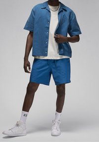 Uomo che indossa una camicia a maniche corte blu con bottoni, pantaloni corti blu abbinati, una t-shirt bianca con grafica, calzini bianchi con il numero "23" e sneakers bianche.