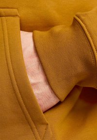 Mosterdgele sweatshirt met een gestructureerde afwerking, voorzien van een buidelzak en geribde manchetten. Close-up van een hand in de zak.