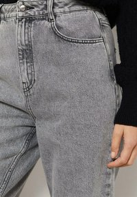 Lähikuva vaaleanharmaista farkuista, joissa on korkeavyötärö ja etutasku, yhdistettynä mustaan pitkähiharaiseen neuleeseen, joka paljastaa oikean käden kyljessä.